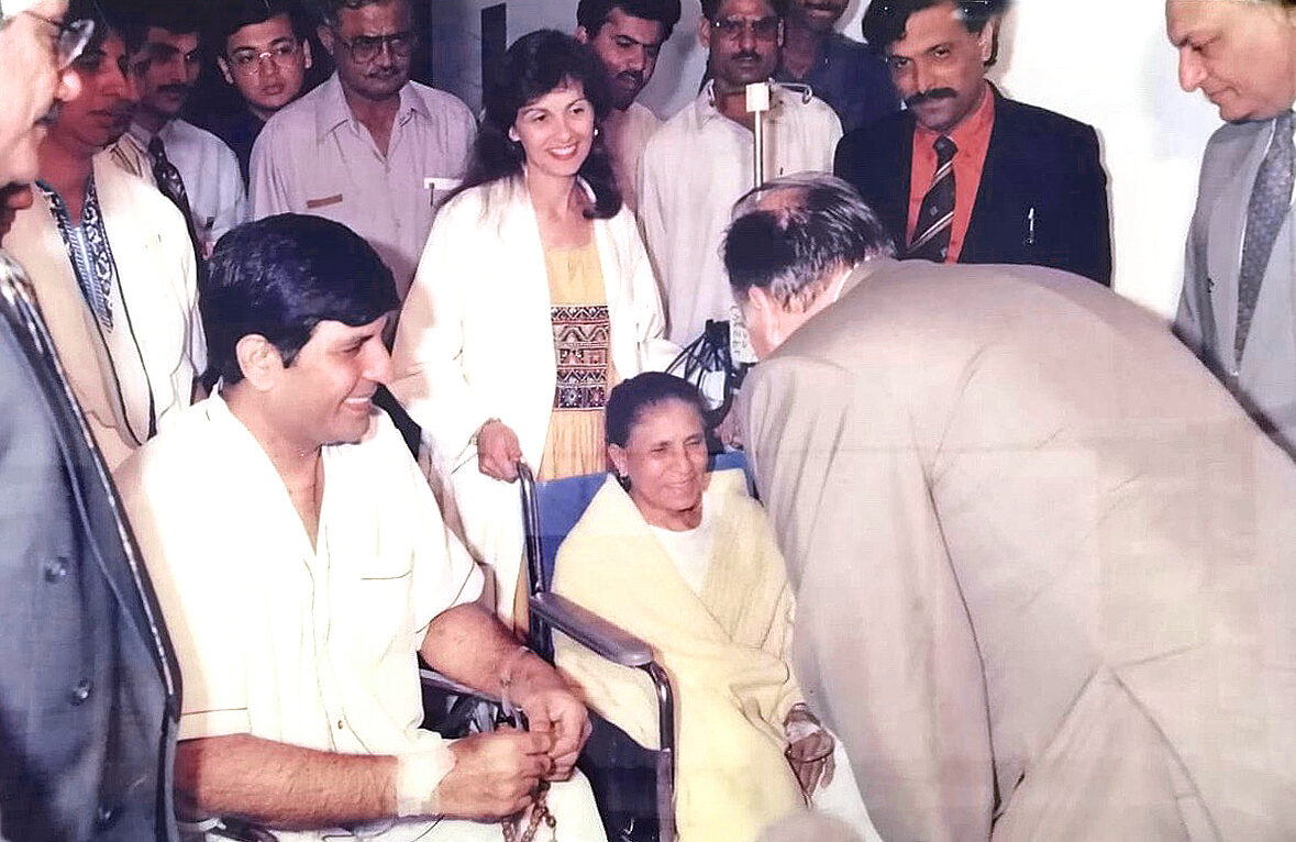 His Highness the Aga Khan bending down to bless the first patient, seated in a wheelchair, with Monique behind her and Dr. Abdulla in a green suit to the left