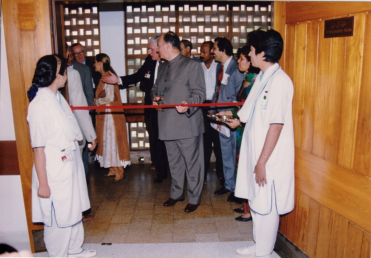 His Highness the Aga Khan holding the ribbon and scissors, not yet cutting, as Princess Zahra Aga Khan and the gathered party turn to look for Dr. Abdulla