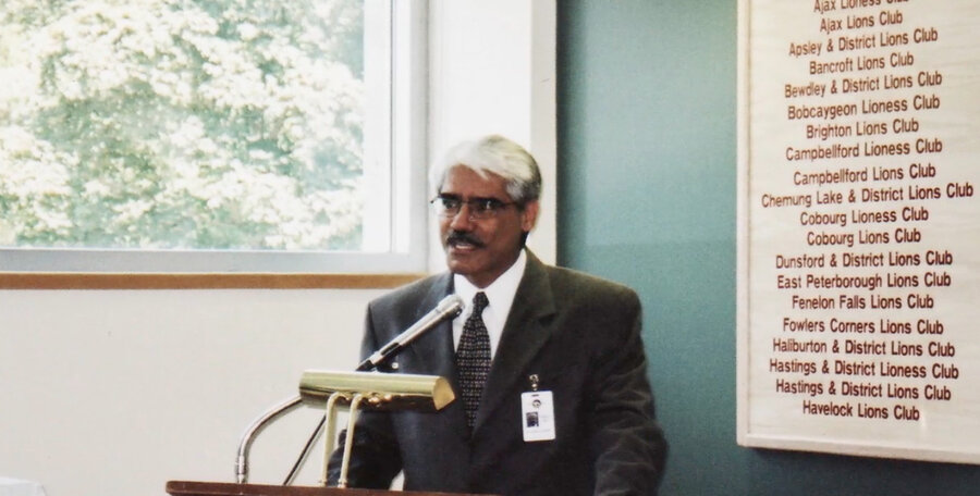 Dr. Abdulla delivering the keynote address at a regional Lions Clubs meeting in Ontario, with a board listing the member clubs behind him